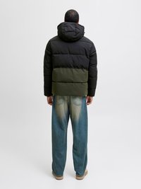 Puffjakke med svart overdel og olivengrønn underdel, har hette og quiltet design. Båret med vide blå jeans og beige sko.