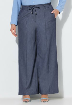 Vrouw draagt blauwe broek met wijde pijpen en een aantrekkoord in de taille, gecombineerd met lichtblauwe sandalen met open tenen en een lichtblauw langermig overhemd.