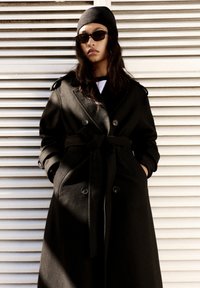 Crni vuneni trench kaput s dvostrukim dugmadi, remenom i džepovima. Model nosi sunčane naočale i crnu kapu. Vertikalna prugasta pozadina.