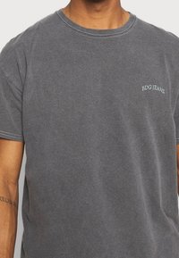 Harmaa puuvillapaita lyhyillä hihoilla, pyöreä kaula-aukko ja pieni vaaleansiniharmaa "BDG JEANS" -logo vasemmassa yläkulmassa.