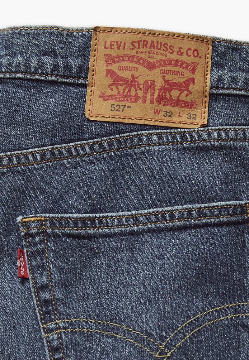 Ciemnoniebieskie dżinsy z denimu z brązową skórzaną łatką z czerwonym i złotym tekstem oraz grafikami. Zawierają małą czerwono logo metkę na tylnej kieszeni.