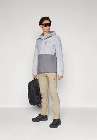 Lys grå og mørk grå vanntett jakke med hette, brukt over en lys blå skjorte. Beige bukser og svarte sko fullfører antrekket. Svart ryggsekk holdt i høyre hånd.