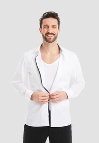White button-up shirt with a contrasting navy trim along the inner placket, long sleeves, and a standard collar, worn over a white tank top.