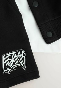 Manga negra con el logo "AllSaints" en blanco en fuente estilizada, descansando sobre una tela blanca con botones a presión negros visibles.