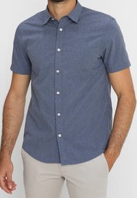 Camicia a maniche corte con bottoni in tessuto navy strutturato, colletto a punta e bottoni bianchi, abbinata a pantaloni chiari.