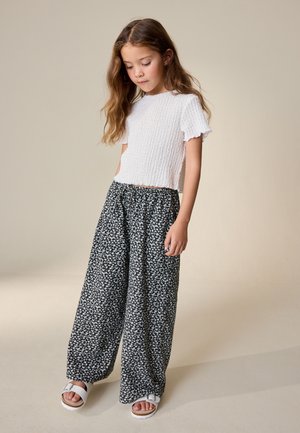 Dziewczyna z długimi brązowymi włosami, ubrana w biały teksturowany krótki top, czarne szerokie spodnie w kwiaty i białe sandały, stojąca z lekko opuszczoną głową.