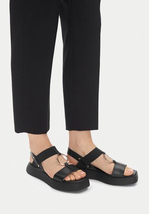 Czarne skórzane sandały na platformie z szerokim paskiem, okrągłym ozdobnym elementem i elastyczną opaską. W połączeniu z czarnymi, krótkimi spodniami.