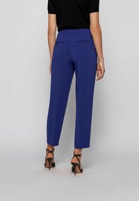 Pantalons ajustés bleu avec une taille haute et deux poches arrière. Portés avec des talons noirs à lanières. Texture lisse, longueur raccourcie.