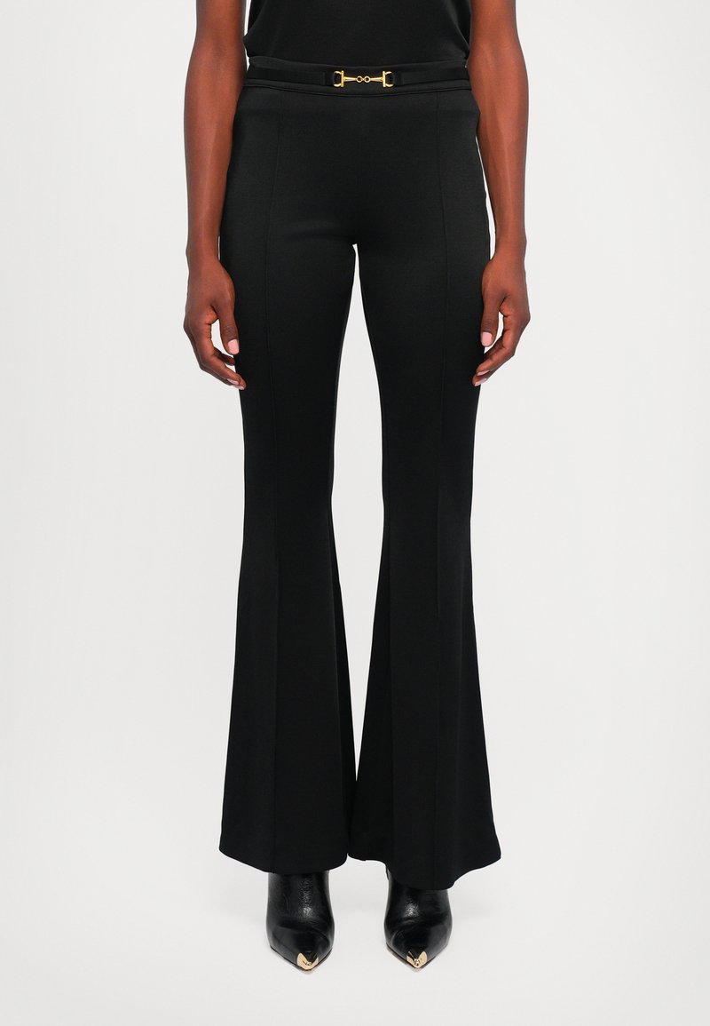 Femme portant un pantalon évasé noir à taille haute et ajusté, avec une ceinture à boucle dorée et des bottines noires à bout pointu.