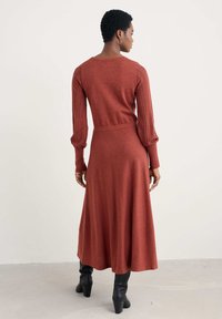 Abito lungo color ruggine lavorato a maglia con dettagli intricati sulle maniche e vita aderente, abbinato a stivali neri con tacco.