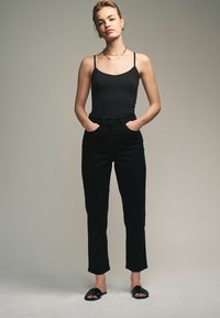 Černý top na ramínka s tenkými popruhy v kombinaci s černými kalhotami s vysokým pasem. Modelka stojí s rukama v kapsách, na nohách má černé ploché sandály.