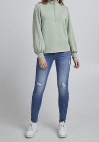 Jersey de color verde menta con cuello alto y cremallera, combinado con vaqueros ajustados azules con detalles desgastados y zapatillas blancas.