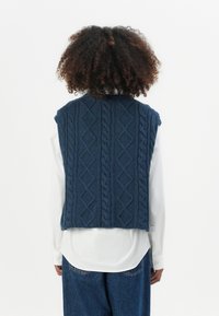 Marineblaue Strickweste mit einem Kabelmuster, hoher Halslinie und ärmellosem Design, getragen über einem weißen Hemd. Strukturierte Oberfläche.