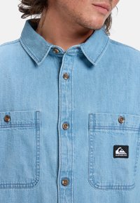 Lichtblauw denim overhemd met een button-down kraag, twee frontzakken, bruine knopen en een zwart Quiksilver logo patch.