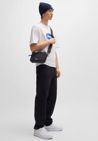 Bolso cruzado negro con textura suave y un logo azul, llevado sobre una camiseta blanca, combinado con pantalones negros y zapatillas blancas.
