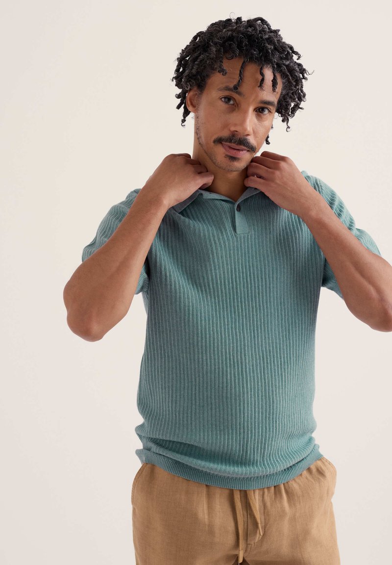 Homme aux cheveux bouclés ajuste le col d'un polo turquoise côtelé à manches courtes, portant un pantalon beige, sur un fond clair uni.