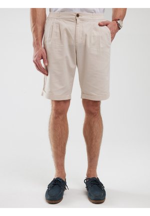 Beige shorts made of lightweight fabric, featuring pleats, a button closure, and rolled cuffs, paired with dark blue casual shoes.