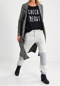 Un cárdigan largo y gris, texturizado, sobre una camiseta negra con texto blanco, unos jeans bloqueados en gris y azul, y botas negras hasta el tobillo.