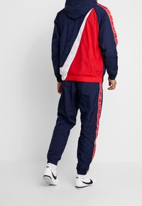 Röd, marinblå och vit träningsdräkt med huva; jackan har snedställda färgblock, elastisk midja och muddar. Bärs med vita sneakers.