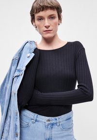 Μαύρο ριμπ μπλουζάκι με μακριά μανίκια, φορεμένο με ελαφριά τζιν μπουφάν. Η μπλούζα έχει φαρδύ ντεκολτέ, ενώ το μπουφάν διαθέτει κουμπωτές λεπτομέρειες.