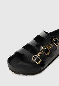 Sandale noire à bout ouvert avec trois brides brillantes et boucles dorées sur fond blanc.