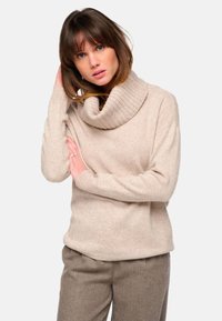 Beiger Pullover mit Schalkragen aus weichem, strukturiertem Stoff, mit gerippten Details und lässiger Passform. Kombiniert mit braunen karierten Hosen.