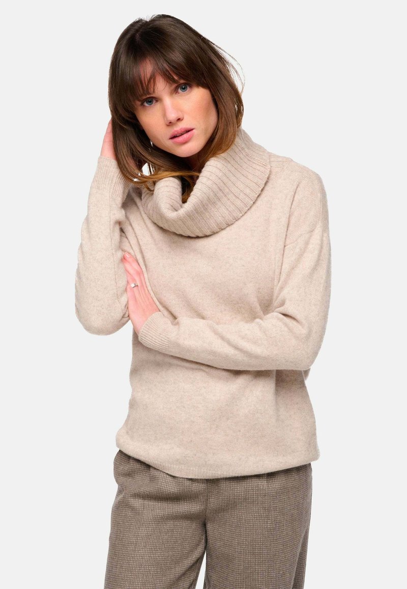 Beiger Pullover mit Schalkragen aus weichem, strukturiertem Stoff, mit gerippten Details und lässiger Passform. Kombiniert mit braunen karierten Hosen.