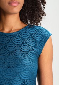 Teal spetsblus med ett vågmönster, kort ärm och en slät yta. Nära bild av halsringningen och axelområdet.