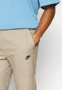 Beige texturerade byxor med elastisk midja och sidofickor, med en svart Nike-logotyp på låret. Kombinerade med en blå t-shirt med korta ärmar.