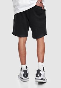 Schwarze Baumwollshorts mit glatter Textur, elastischem Bund und seitlichen Taschen. Kombiniert mit weißen und schwarzen Sportschuhen.