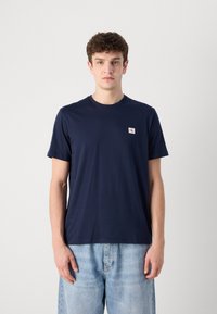 Marineblaues Baumwoll-T-Shirt mit Rundhalsausschnitt, kurzen Ärmeln und einem kleinen CK-Logo-Patch auf der linken Brust. Genossen mit hellblauen Jeans.