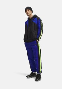 Sportieve trainingsset in blauw en zwart met neon groene strepen. Lichtgewicht materiaal, ritssluitingjack, bijpassende broek en zwarte sneakers.