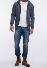 Uomo in t-shirt bianca, felpa con cappuccio grigia scura e zip, jeans blu e stivali in pelle marrone con lacci, in piedi contro uno sfondo bianco.