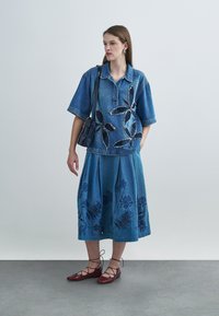 Chemise en jean avec broderie florale, associée à une jupe plissée en jean ornée de motifs floraux sombres. Ballerines rouges et petit sac noir.