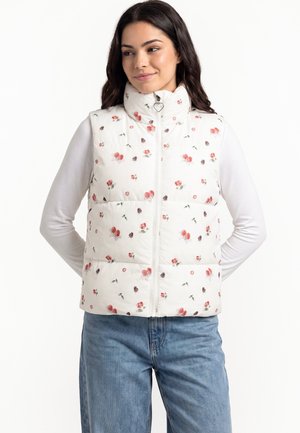 Jonge vrouw die een wit gevoerd vest met bloemmotief en hartvormige ritstrekker draagt, een witte top met lange mouwen en een blauwe spijkerbroek.