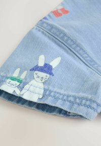 Svetlo modra denim tkanina prikazuje dva risana zajčka z vezenimi klobuki, enega zelene in enega vijolične barve, blizu všitega roba.