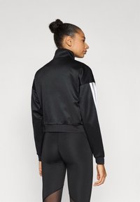 Črna satenasta jakna z visokim ovratnikom in belimi poudarki na ramenih, z oprijetim spodnjim delom in gladko teksturo. Oprijete črne legice v kombinaciji.