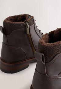 Bottes basses en cuir marron avec une doublure intérieure marron texturée, un revers à motif chevrons contrastant, fermeture à lacets et fermeture éclair latérale.