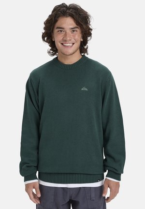 Suéter de punto verde con cuello y puños acanalados. Presenta un pequeño logo en el pecho y un look en capas con una camisa blanca debajo.