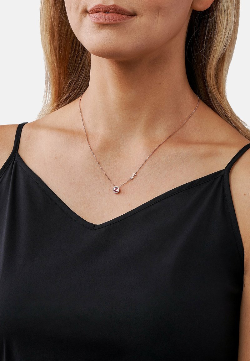 Femme portant un délicat collier en or rose avec un pendentif en pierre précieuse rose ronde sur une fine chaîne, vêtue d'un haut noir sans manches.