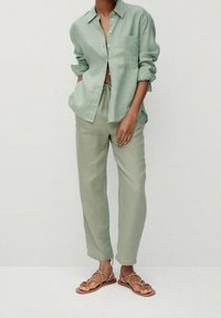 Lichtgroen linnen overhemd met opgerolde mouwen, gecombineerd met bijpassende loszittende linnen broek en platte sandalen met bandjes op een neutrale achtergrond.
