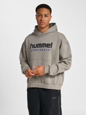 Mand iført taupe hummel sportswear hættetrøje og sorte hummel sportswear bukser, stående med hænderne foldet på en hvid baggrund.