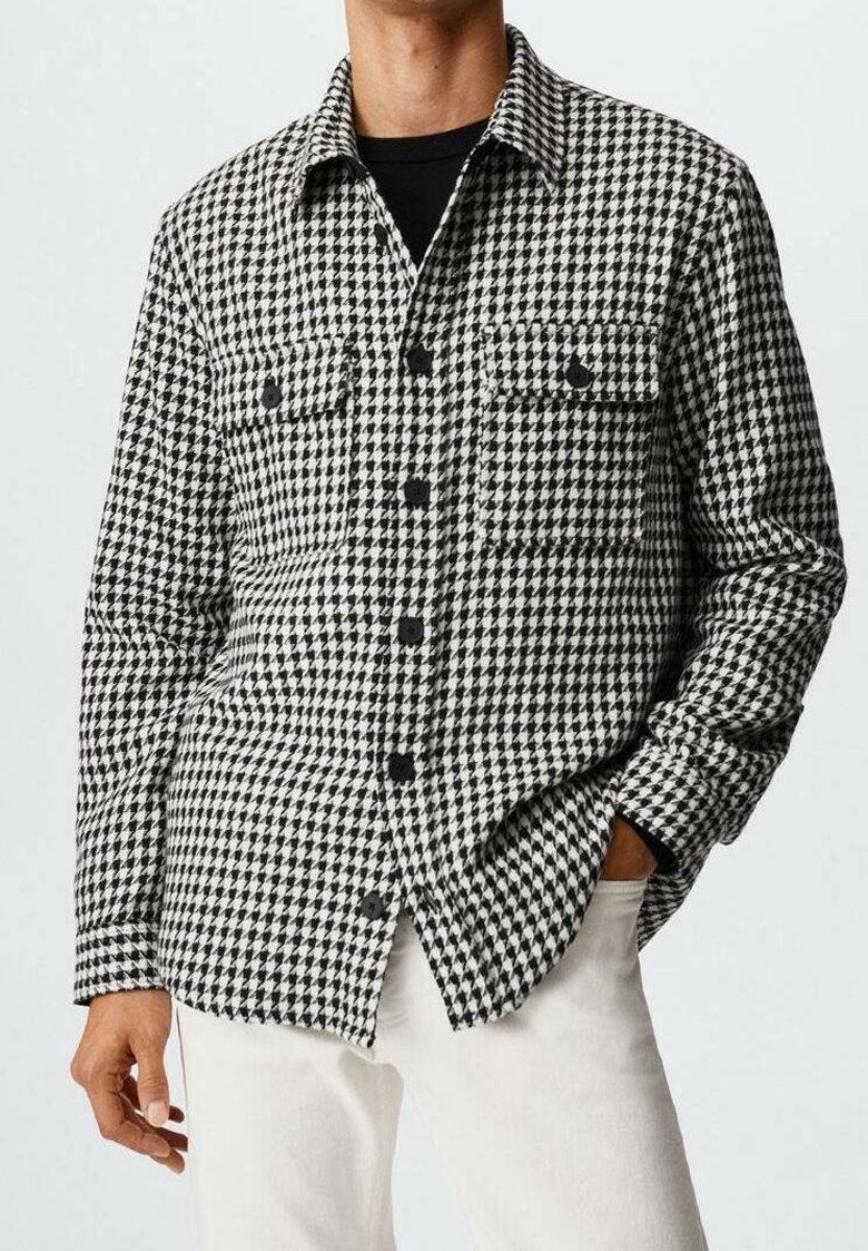 Homme portant une chemise à boutons à motif pied-de-poule noir et blanc avec des poches poitrine, associée à un pantalon blanc. Une main dans la poche.
