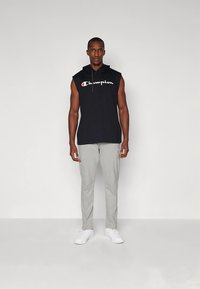 Juodas be rankovių gobtuvas su "Champion" logotipu, derinamas su šviesiai pilkomis sportinėmis kelnėmis ir baltais sportbačiais. Paprasta, kasdieninė sportinė apranga.