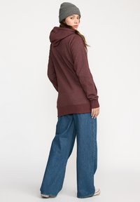 Un sweatshirt bordeaux avec une capuche à cordon, des manches longues et un ourlet côtelé, associé à un pantalon en denim bleu à jambes larges et un bonnet en tricot gris.