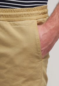 En vuxenhand som vilar i framlommen på beiga byxor med resårmidja, iklädd en marinblå och vit randig skjorta.