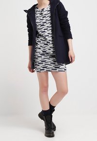 Personne portant des bottes noires, des chaussettes bleu marine, une veste zippée bleu marine et une robe mi-longue à motifs géométriques noirs et blancs.