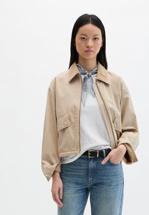 Jeune femme aux longs cheveux noirs portant une veste courte beige, un T-shirt blanc, un jean bleu, une ceinture noire et un foulard bleu clair à motifs noué autour du cou.