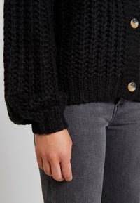 Svart stickad cardigan med en texturerad mönster, med stora knappar och uppvikta ärmar, ihop med grå jeans. Närbild av �ärmen.
