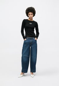 Camisola preta de manga comprida com logótipo, combinada com jeans azuis de perna larga. Sapatos de salto altos brancos com bico pontiagudo completam o visual. Design simples e casual.